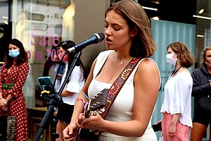 A teenage girl plays guitar by a microphone on a street