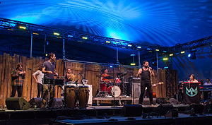 Eight men performing on a stage. From left to right; two horns, timbales, shekere, drums, vocals, bass (obscured), keyboards.