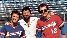 Three men standing in front of blue stadium seats. One is wearing a blue baseball jersey, one is in a white dress shirt, and one is in a red baseball jersey.