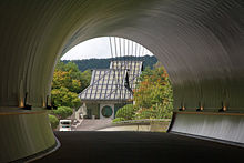 A passageway at the Miho Museum. A curving circular tunnel opens to reveal a building with a tall sloping roof and a circular window in the front door.