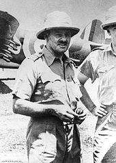 Portrait of a man in tropical military uniform. He is wearing a bucket hat, and military aircraft can be seen in the background.