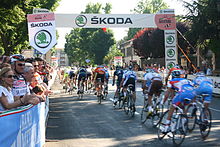 A group of cyclists ride under an arch erected on the road. Spectators watch at the roadside.