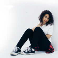 Alessia Cara in a white top and black pants afront a white background.