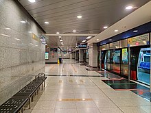 A train arrives at the CCL platform (on the right). On the left is a grey wall with a black rectangular seat engraved with the station's name.