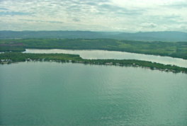 A panorama of a lake with a thin green strip of land and homes that reflect on the lake's edge, followed by a small area of lake waters, a larger stretch of green land, a mountain range and a cloudy sky overhead.