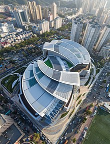 A photo of Olympia 66, a large retail building designed to resemble the Chinese twin carp emblem, a symbol of wealth and abundance in Chinese culture. The building is primary white in color with open green space and spiraling layers of gray.