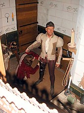 A wax figure of a man stands, preparing for battle. The figure sports a bowl cut and wears a khaki doublet and dark brown breeches. At its legs is the wax figure of a kneeling squire.