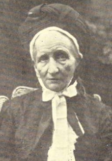 An older white woman, wearing a substantial bonnet or headwrap, tied under her chin, and a dark dress with a white ruffle down the chest.