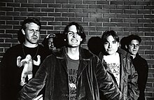 A black and white photograph of five members of the group Pavement standing in front of a brick wall