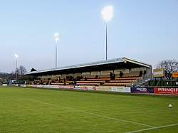 A single-tiered stand which has nine rows of alternating gold and black seats. In front of the stand is a field of grass upon which a solitary football sits