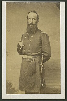 A standing bearded man in civil war uniform, sword at his side and right hand tucked in his jacket while left hand is behind his back. Posed for this photograph eyes looking at the camera and body slightly off square to the right from facing the camera.