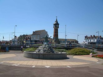 A fountain with a sculpture of a person; a clock tower, people and buildings are in the background