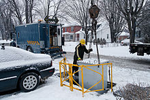 A man with a hard hat helps a machinery operator lower a transformer to an underground pit in a residential neighborhood. The roads and cars are covered with snow.