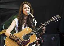 A woman is on stage singing into a mic and playing a guitar.