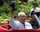 An elderly almost-bald man wearing a striped button-up shirt and a tie adorned with a pattern of baseballs waves a cap from a convertible.