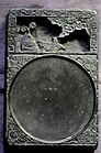 A white, rectangular stone tablet with two sections. The top quarter section contains a carving of a fish, and the bottom three-fourths contain a shallow, circular indent for the ink.