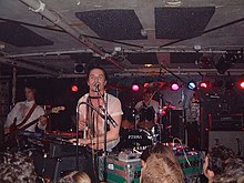 A band play in a crowded room. A man is playing keyboards while singing into a microphone; behind him is a drummer and to one side is a guitar player.