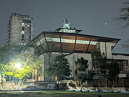 Nighttime photo of a large, square concrete building topped by an inverted trapezoidal section (wider at the top than the bottom) ringed by a row of windows, and a much smaller, almost all-glass rectangular room atop that, in turned topped with a narrow glass pyramid partly surrounded by decorative metal bars. In the foreground are several trees and a sports field, and in the background to the left there's a cold gray, rectangular high-rise office building