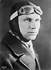 Profile of a white man wearing a thick coat and tight-fitting leather aviator's cap, strapped under the chin, with goggles pushed up onto his forehead.