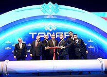 Seven middle aged men in dark suits stand behind a large white horizontal pipe. There is a large TANAP logo behind them and six of them are holding the large red valve wheel protruding up from the pipe