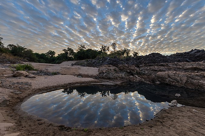 图为日出时分，在老挝四千岛东阔岛一片带岩石的沙滩上，反射在池塘水面上的绳状灰云和白云。