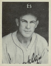 A man in a white baseball jersey and a dark cap with a white "B" on the front looks toward the camera.