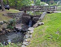 A wooden bridge over a small dam over which water falls into a stream flowing out of the picture at lower left. Behind it is a large, still pond; to the left a black sport-utility vehicle is parked