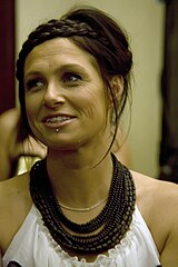 The 32-year-old Chambers wears a white dress with bare shoulders. She has numerous brown necklaces and a silver one. She has a chin stud below her lower lip and is looking to her right with a smile as she speaks.
