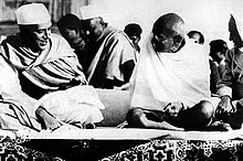 Two smiling men in robes sitting on the ground, with bodies facing the viewer and with heads turned toward each other. The younger wears a white Nehru cap; the elder is bald and wears glasses. A half dozen other people are in the background.
