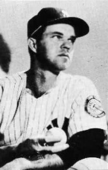 A man in a white baseball uniform and dark cap holding a baseball in his right hand.