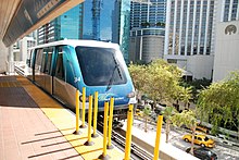 A people mover vehicle is pulling into an elevated metro station with large buildings in the background.