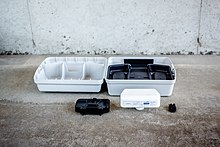 A white box that looks like a typical rat bait station sitting on a concrete floor opened up to show the interior, with a white cartridge with the ContraPest bait station branding on it containing the sweet liquid that the rat will drink whilst inside the bait station