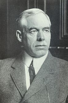 This is a headshot of an older white man with short hair, wearing a double breasted jacket. He has a dark tie on that has a straight pin in it and his jacket has what looks to have a watch chain hanging from one of his lapels.