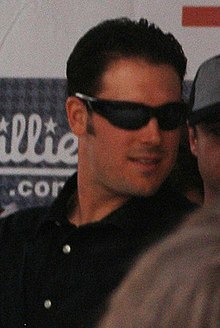 A dark-haired man with sideburns wearing a white shirt and dark sunglasses