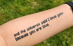 手臂上的纹身，内容为“and the universe said I love you because you are love”，使用了与Minecraft相似的字体。