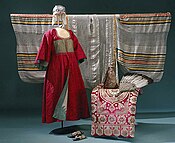 Three pieces of cloth photographed. A red suit extending the lower body is placed on a mannequin at center left, a white cloth with black and gold embroidery is at the back, a red cloth with ornate patterns is at bottom right