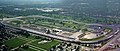 Aerial view of a rounded rectangular raceway that is partially surrounded by grandstands; within the racetrack is a pagoda and golf course