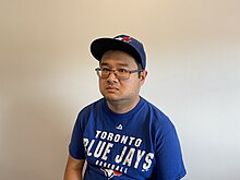 An average-build short-haired slightly tanned East Asian man in his mid-30s with an intentional stubble wearing eyeglasses, an official Toronto Blue Jays T-shirt from Majestic, and an official Toronto Blue Jays baseball cap from New Era Caps
