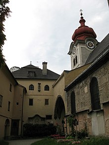 Part of a partially planted courtyard. As viewed, on the western and northern sides is a four-storey building with a smooth yellow render and dark doors and windows; on the eastern side is an archway surrounded with the same render, with the remainder of the building face in stone with dark windows. Above the arch is a square, white clocktower with a red, octagonal, bulbous spire.