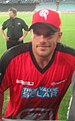 An Australian cricketer wearing a red-black jersey. The cricket field and an another person can be seen in the background.