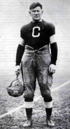 A man wearing a football uniform from the early 20th century. His black shirt has a single large white stripe on each bicep and the letter "C" on the front. He is holding his leather helmet by his side.