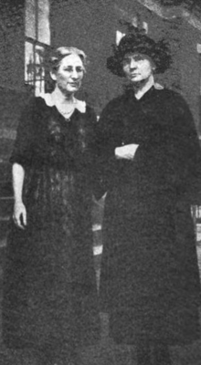 Two white woman standing outdoors, both wearing dark dresses; the woman on the right is also wearing a dark brimmed hat