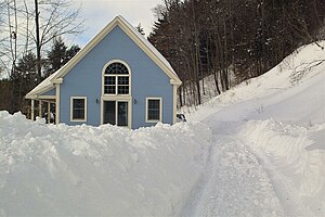 A blue house in a snow-covered property on a hillside. Tire tracks are visible on the bottom-right.