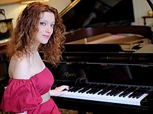 Licia Missori sitting in front of a black Essex grand piano wearing a red dress and looking at the camera.