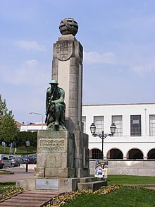 Photograph of a monument that has a bronze soldier bent on one knee holding a rifle, and behind him is a tall pillar with a round shape on the top
