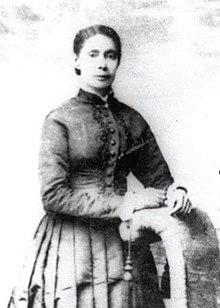 Black and white photograph of a black woman in a Victorian dress with her hands resting on a curved chair back