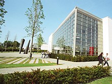 This is the side view of the Nursing Building. It is a glass building with white walls as borders. The picture is taken during the day. There are two or three tall trees in front, with two cone like decorative structures in front. There are two groups of students (groups of 3 and 2 respectively) walking on the pathway. In the background, there are similar looking buildings on the right, and trees on the left. In the foreground, there is a neatly manicured lawn with small hedges.