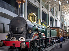 A black, re, and gold-trimmed locomotive with coal tender and two passenger cars sits on a rail siding at the museum