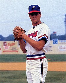 A man wearing a white baseball uniform with blue and red trim and a blue cap with a white "N" on the center stands with his hands held together in his brown leather glove.