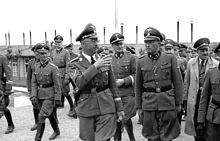 A group of Nazi officers, including Heinrich Himmler, Franz Ziereis, Karl Wolff and August Eigruber shown walking and talking through the camp, with one of the huts in the background.